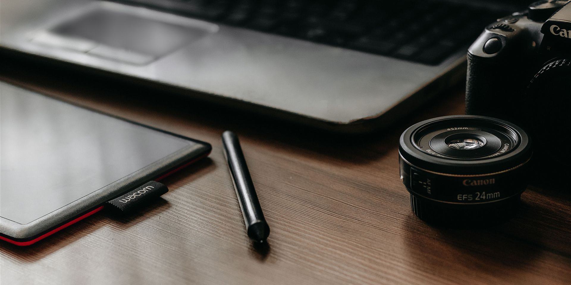 Ein Arbeitsplatz mit Laptop, Tablet, Kamera und Objektiv, ideal für kreative Arbeiten und Fotografie.